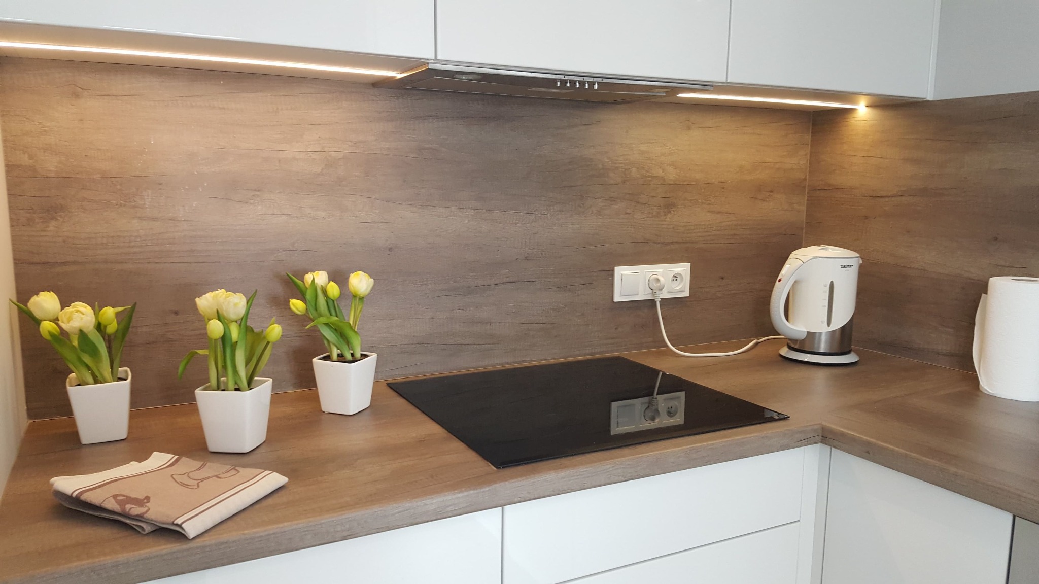 Uporządkowana kuchnia z drewnianym blatem, trzema doniczkami z żółtymi tulipanami i czajnikiem elektrycznym; minimalistyczny styl.