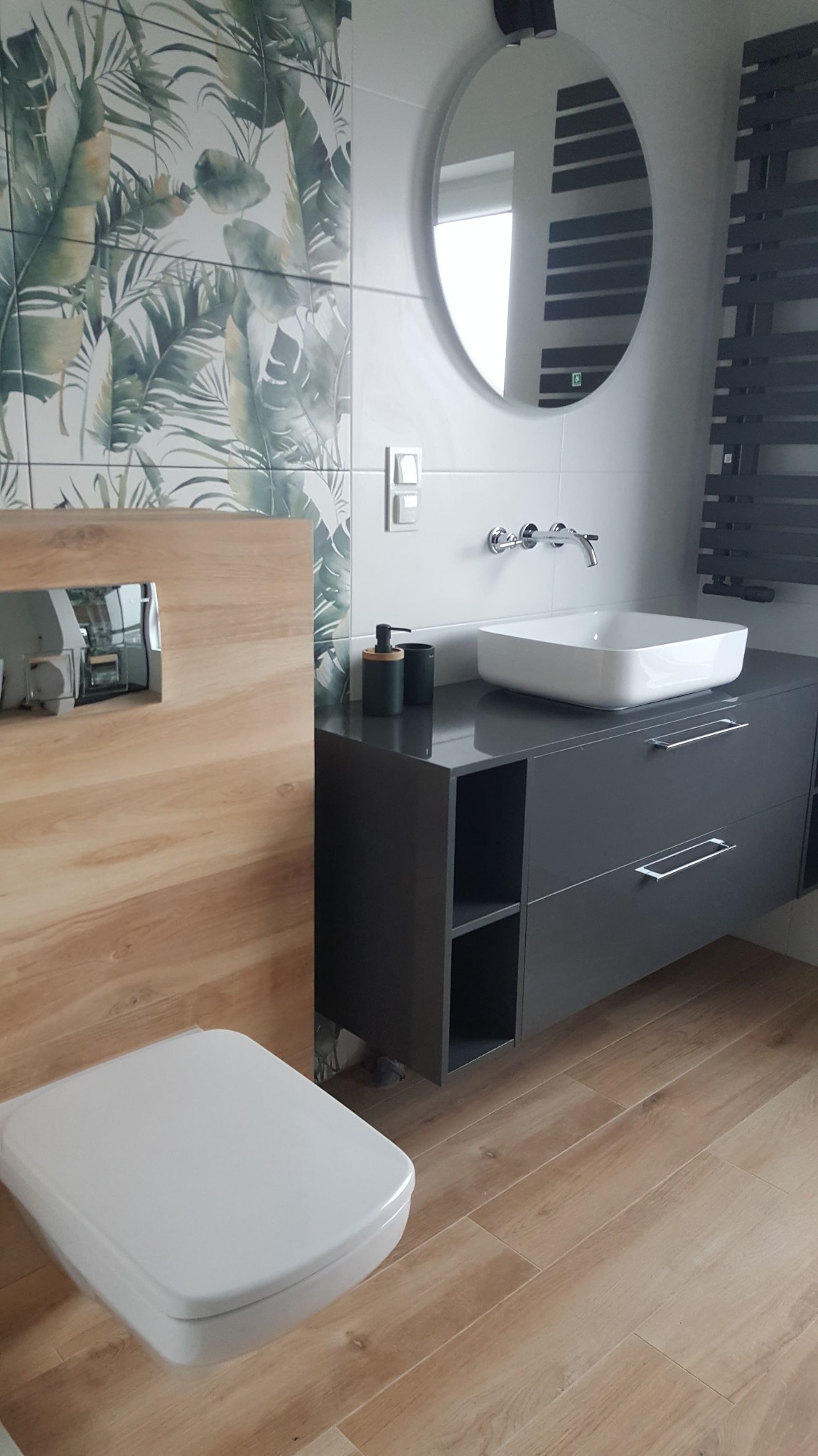 Nowoczesna łazienka z drewnopodobnymi płytkami podłogowymi i ściennymi, umywalką nablatową na szarej szafce z szufladami, okrągłym lustrem z podświetleniem LED oraz toaletą podwieszaną.