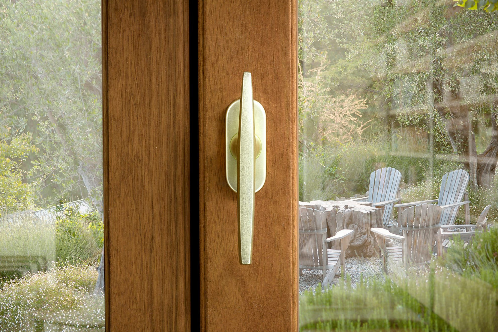 Drewniane okno z zamkniętym, metalowym uchwytem, przez szybę widoczne krzesła ogrodowe i zieleń.