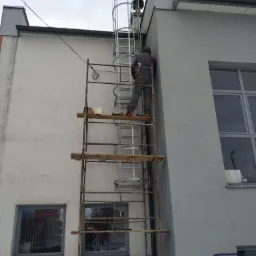 Pracownik na rusztowaniu przy budynku, montujący metalową drabinę ewakuacyjną z koszem ochronnym, widok z dołu na szarą fasadę i pochmurne niebo.