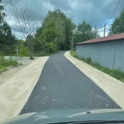 Asfaltowa droga z betonowymi poboczami, otoczona zielenią i budynkiem gospodarczym, widziana z perspektywy pojazdu z pochmurnym niebem w tle.
