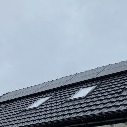 ELEKTRYK-FOTOWOLTAIKA - Czarny dach z dachówkami i oknami dachowymi, na którym zamontowano panele fotowoltaiczne w pochmurny dzień.
