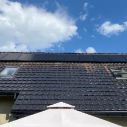 Panele fotowoltaiczne 7.3kW zamontowane na brązowym dachu z dachówki ceramicznej, widoczne dwa okna dachowe i fragment białego parasola ogrodowego na tle błękitnego nieba z chmurami.