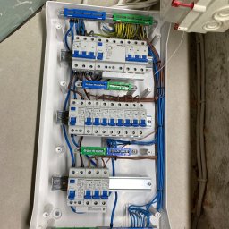 ELEKTRYK-FOTOWOLTAIKA - Rozdzielnica elektryczna z zamontowanymi bezpiecznikami Schrack, widoczne przewody w kolorach niebieskim, brązowym i żółto-zielonym, zamocowane do listew zaciskowych.