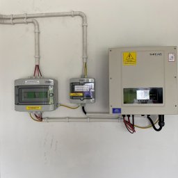 ELEKTRYK-FOTOWOLTAIKA - Zestawienie komponentów instalacji fotowoltaicznej: rozdzielnice DC i AC, inwerter SOFAR z widocznymi przewodami, zamontowane na białej ścianie wewnątrz budynku, z widocznymi rurami ochronnymi...