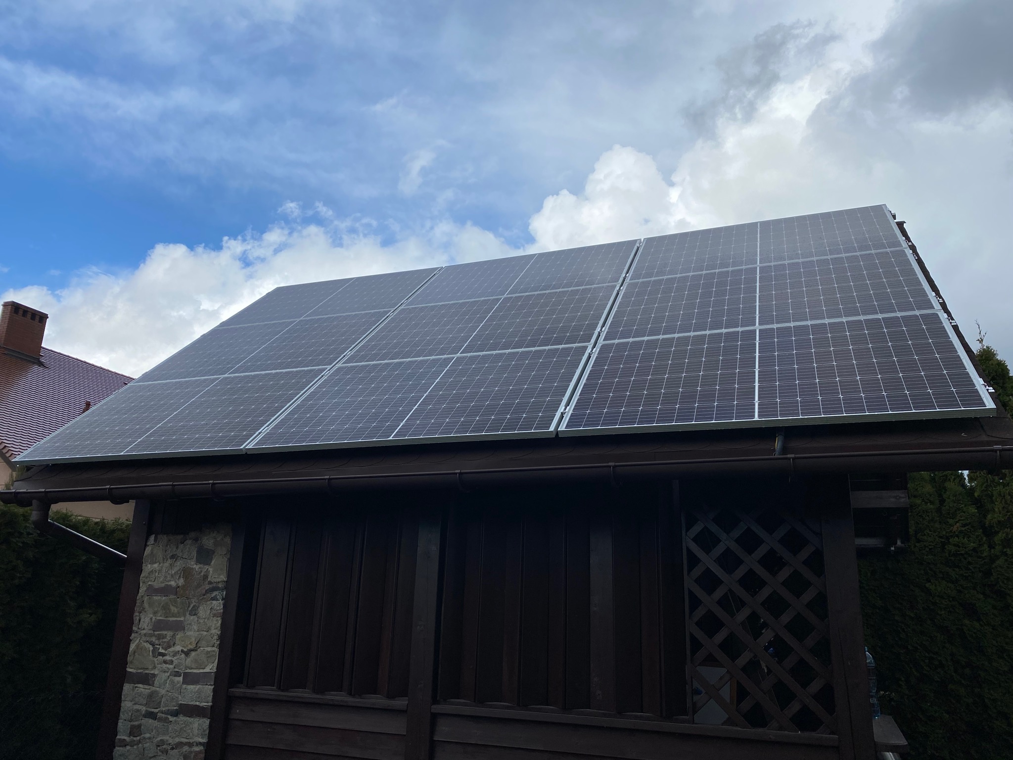 Panele słoneczne zamontowane na dachu drewnianej altany, widok z dołu na tle błękitnego nieba z chmurami.