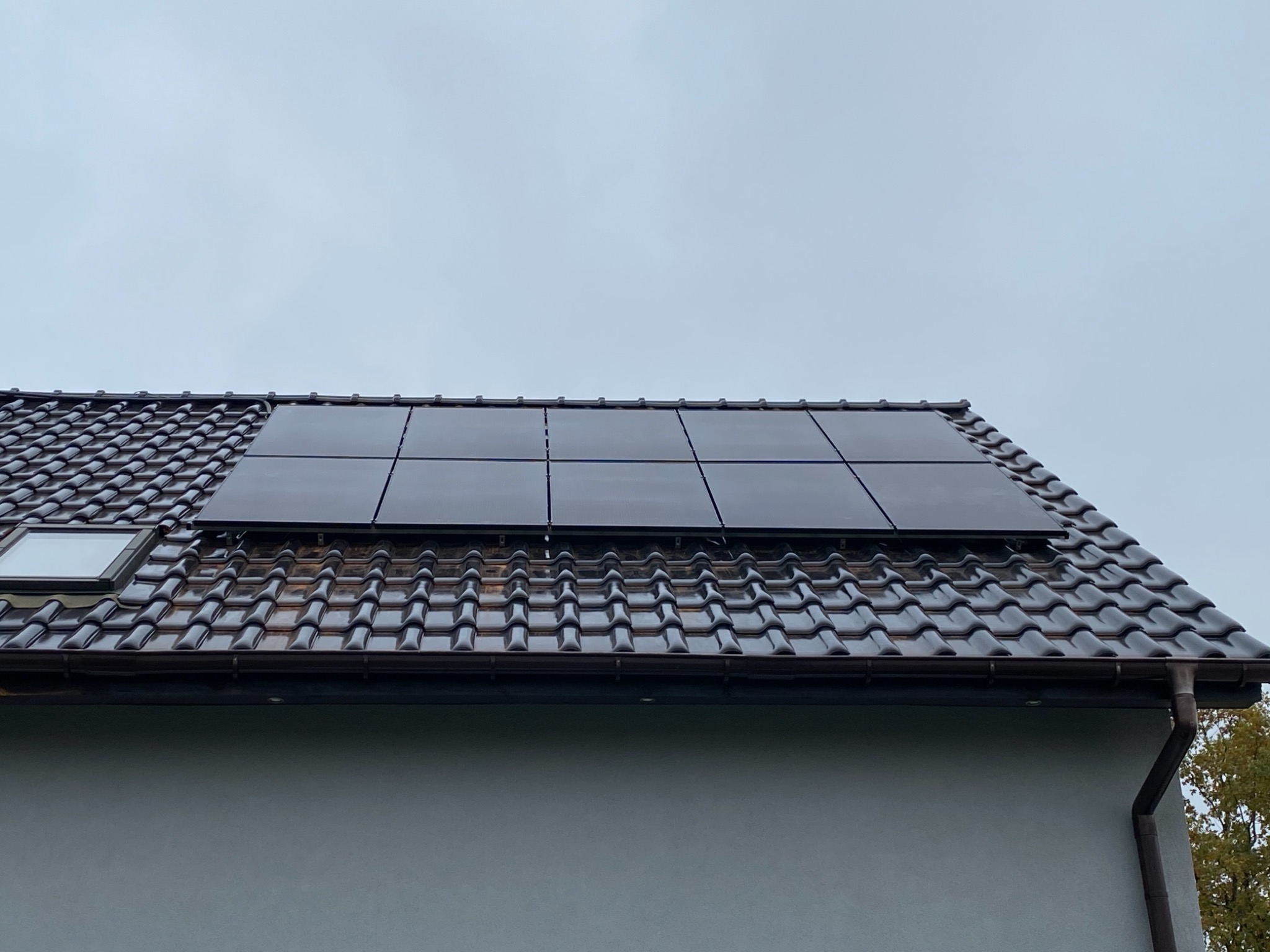Czarne panele słoneczne zamontowane na brązowym dachu z dachówki ceramicznej, widoczne okno dachowe i fragment elewacji budynku w pochmurny dzień.