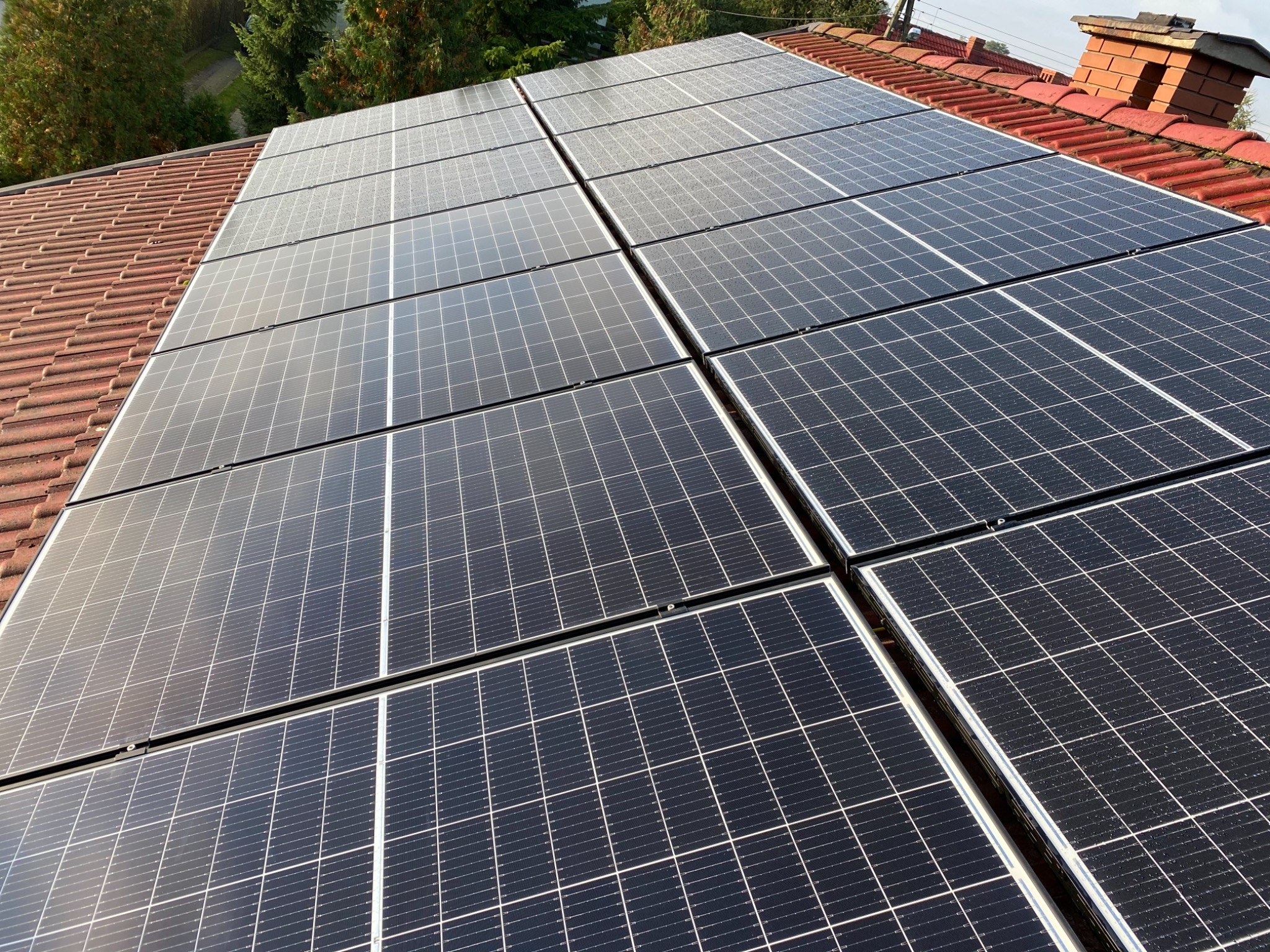 Panel słoneczny na dachu z czerwonej dachówki, widok z góry, lekko pochmurne niebo.