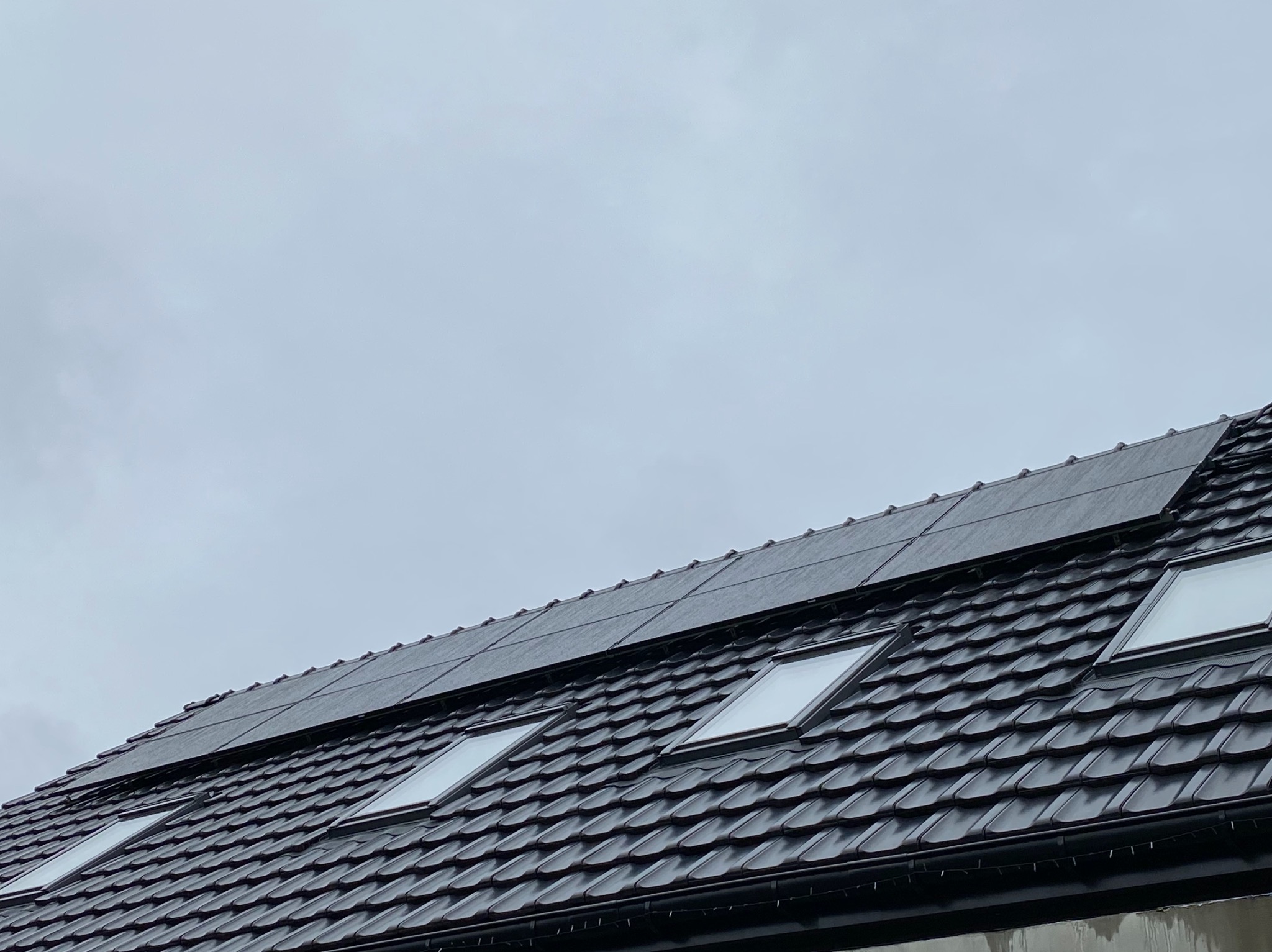 Czarny dach z dachówkami i oknami dachowymi, na którym zamontowano panele fotowoltaiczne w pochmurny dzień.