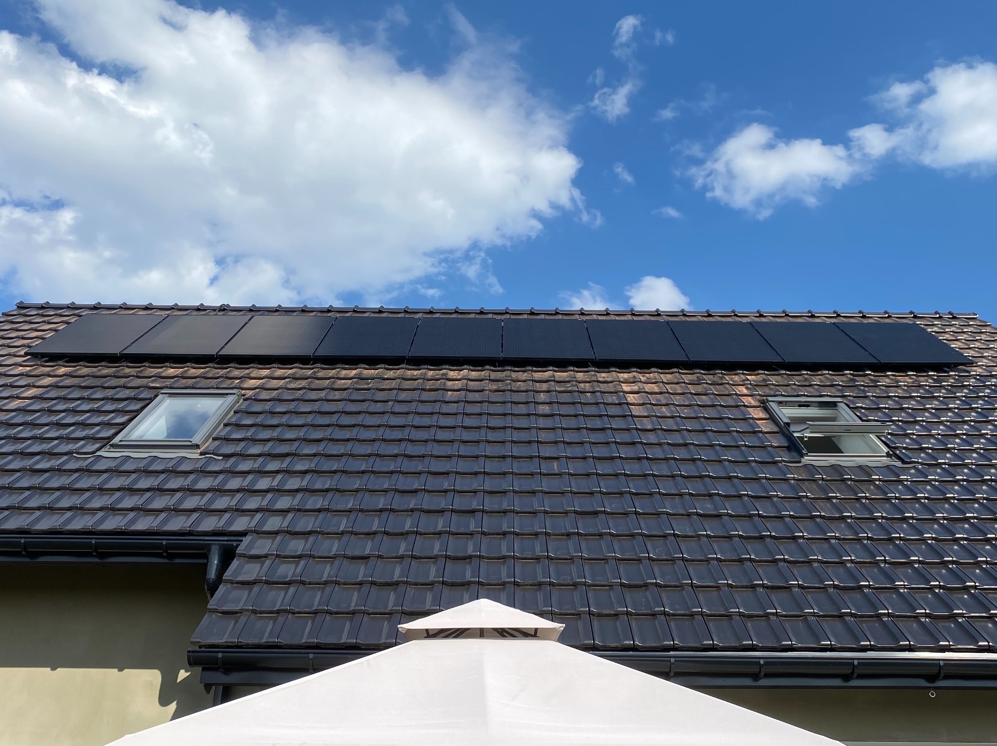 Panele fotowoltaiczne 7.3kW zamontowane na brązowym dachu z dachówki ceramicznej, widoczne dwa okna dachowe i fragment białego parasola ogrodowego na tle błękitnego nieba z chmurami.