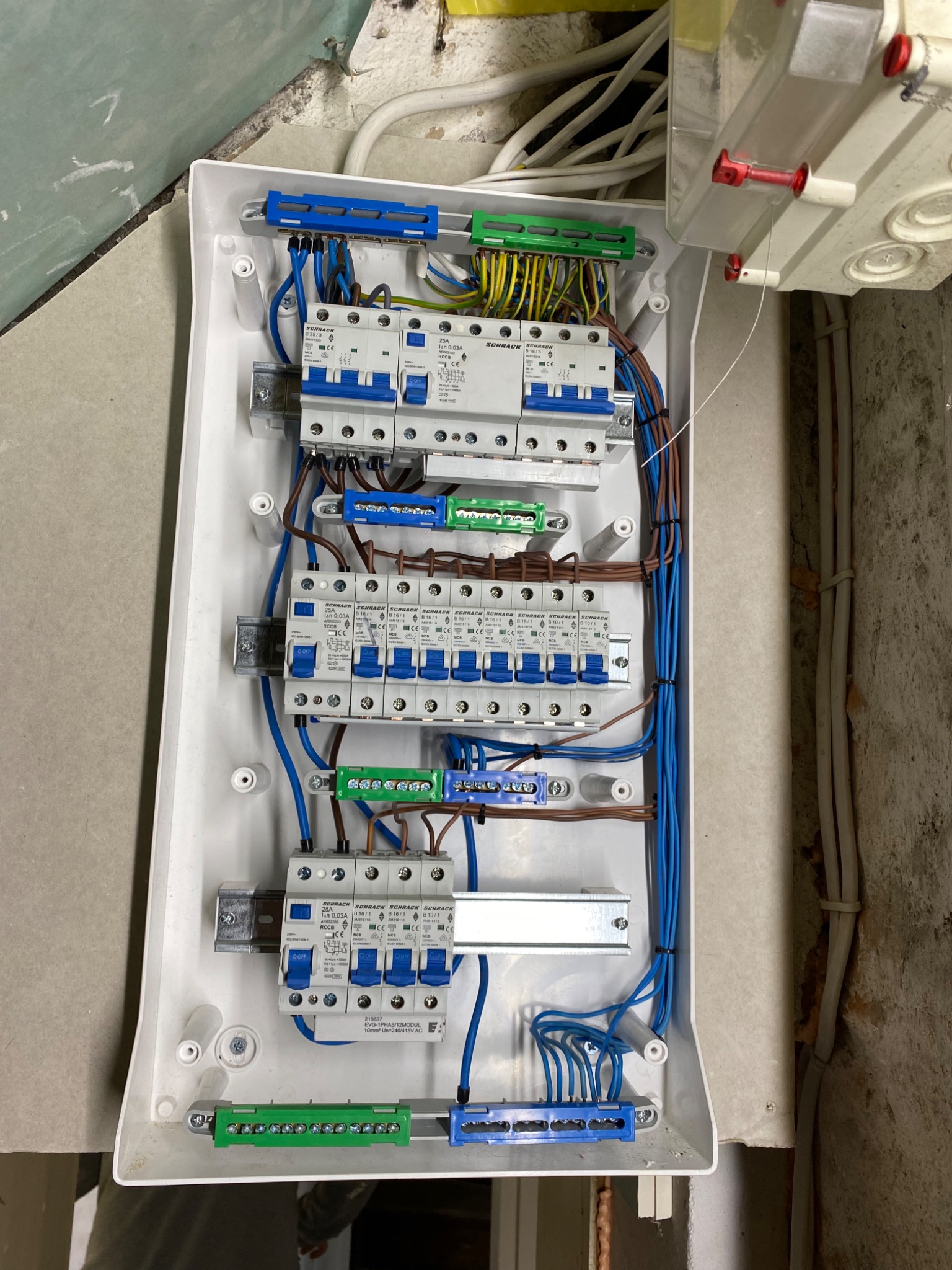 Rozdzielnica elektryczna z zamontowanymi bezpiecznikami Schrack, widoczne przewody w kolorach niebieskim, brązowym i żółto-zielonym, zamocowane do listew zaciskowych.