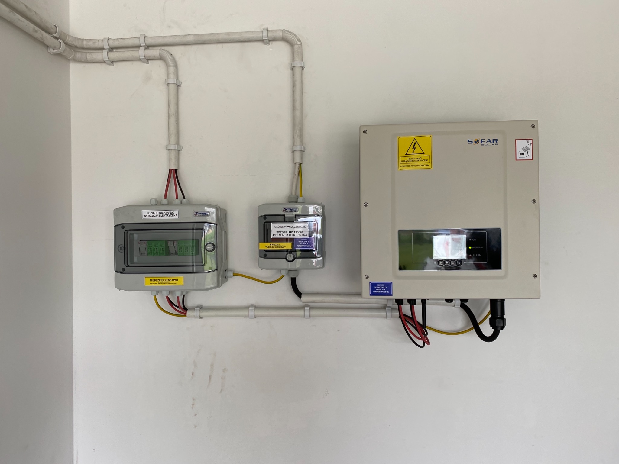Zestawienie komponentów instalacji fotowoltaicznej: rozdzielnice DC i AC, inwerter SOFAR z widocznymi przewodami, zamontowane na białej ścianie wewnątrz budynku, z widocznymi rurami ochronnymi...