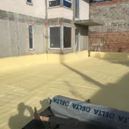 Żółta izolacja natryskowa pokrywająca płaski dach budynku w trakcie budowy, widoczny fragment rolki folii Delta i cień osoby.