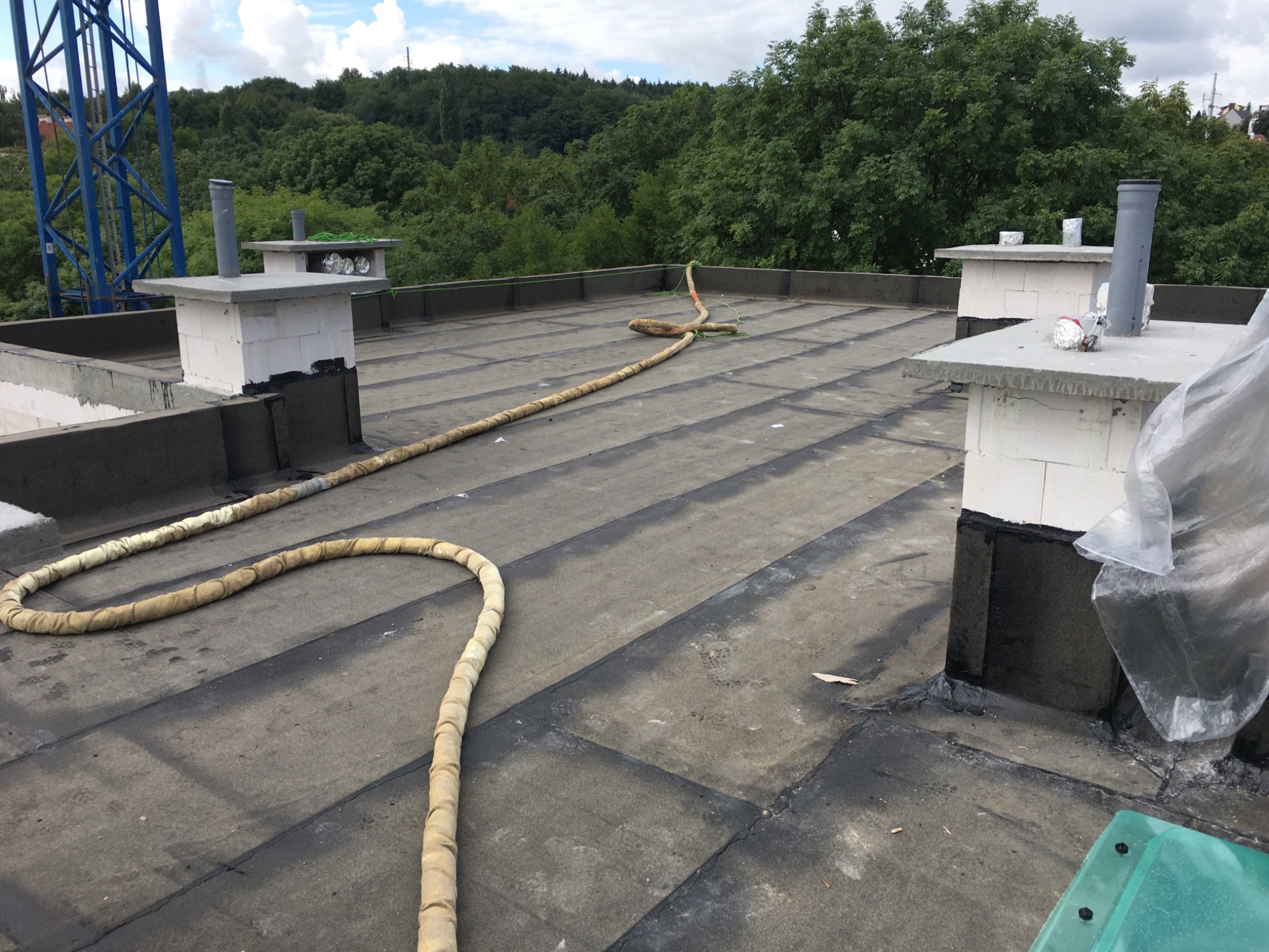 Widok z góry na płaski dach pokryty papą, z widocznymi kominami, zabezpieczeniami i fragmentami zieleni w tle.