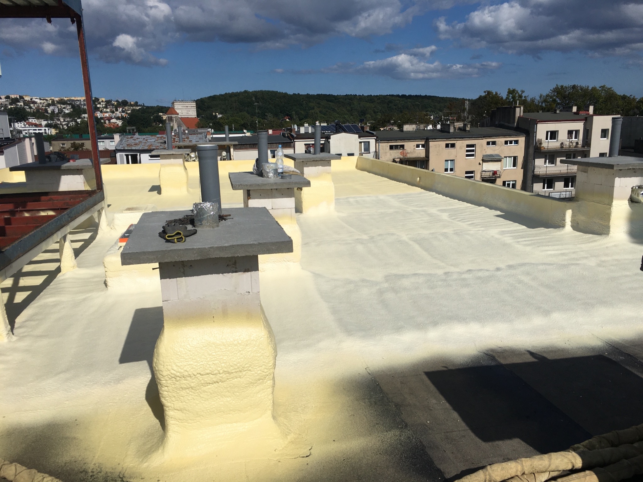 Jasna, świeżo natryśnięta izolacja na płaskim dachu budynku, z widocznymi kominami i narzędziami, w tle zabudowa miejska i zielone wzgórza.