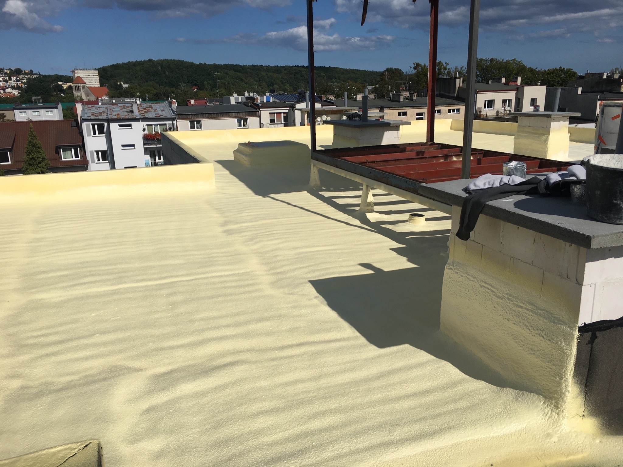 Jasnożółta warstwa izolacyjna na dachu budynku, widoczne elementy konstrukcji stalowej i kominy, w tle zabudowa miejska i zieleń.
