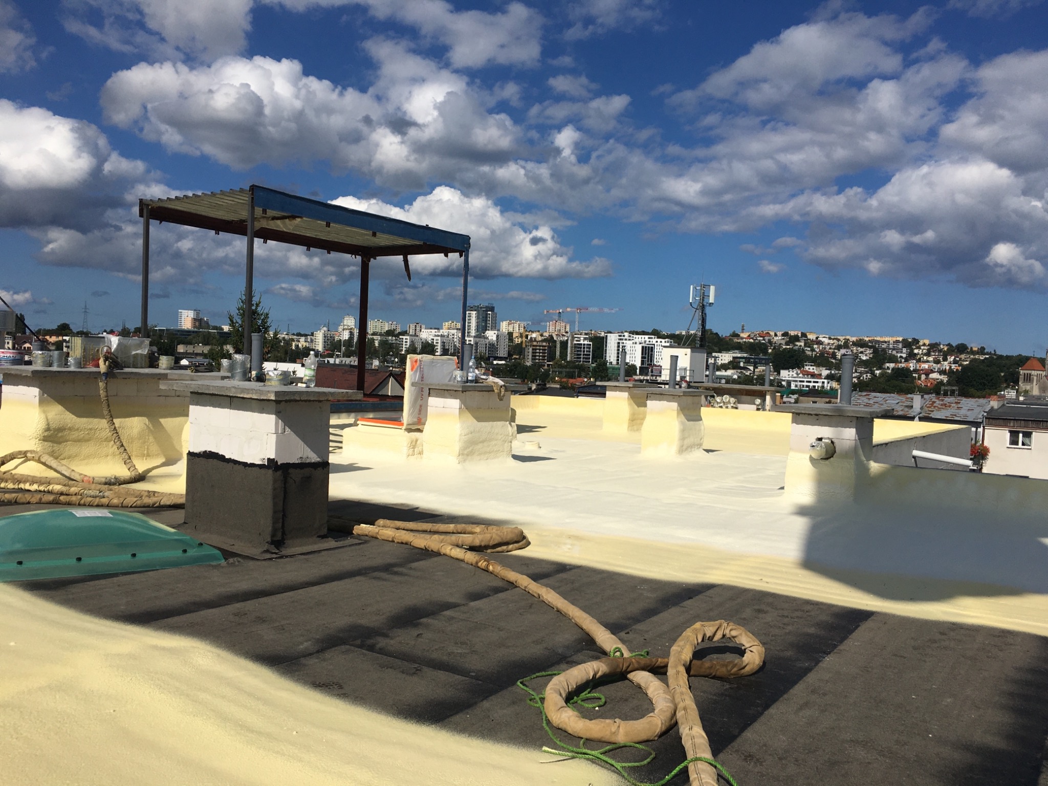 Jasnożółta warstwa izolacyjna na dachu budynku z widocznymi elementami infrastruktury, w tle panorama miasta pod błękitnym niebem z chmurami.