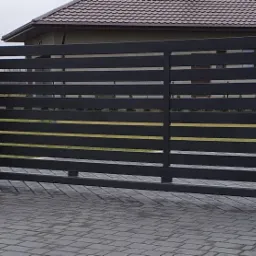 Nowoczesna, przesuwna brama wjazdowa z poziomych, czarnych profili, osadzona pomiędzy białymi słupkami i jednym słupkiem z imitacji drewna, na tle kostki brukowej i fragmentu domu.