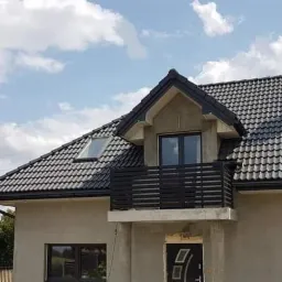 Surowy stan elewacji domu jednorodzinnego z ciemną dachówką i czarną, minimalistyczną balustradą balkonową, widok z zewnątrz w słoneczny dzień.