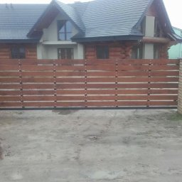 Mobilne Usługi Stolarsko - Spawalnicze Sylwester Kostecki - Przesuwna brama z poziomych desek w kolorze brązowym, zamontowana między murowanymi słupkami z cegły klinkierowej, na tle drewnianego domu z ciemnym dachem.