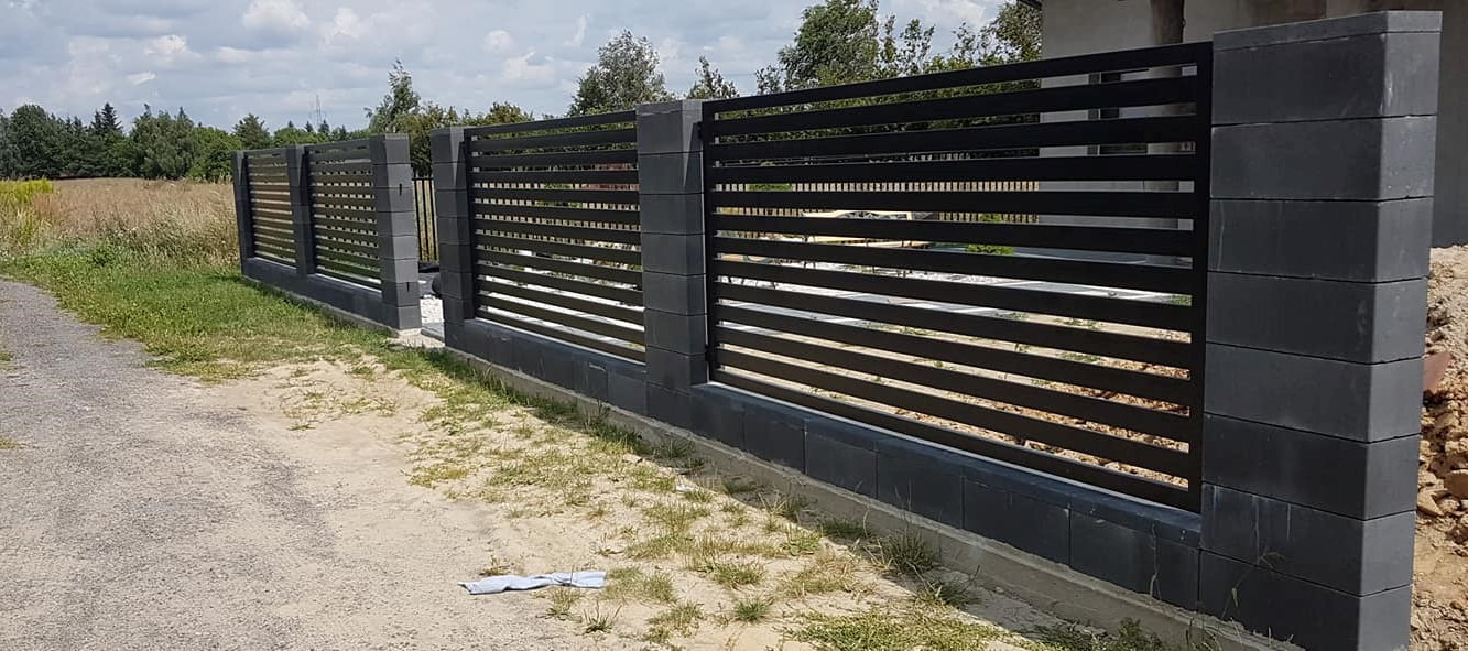 Nowoczesne ogrodzenie panelowe z poziomych, czarnych profili stalowych osadzonych na szarych, betonowych słupkach i podmurówce.