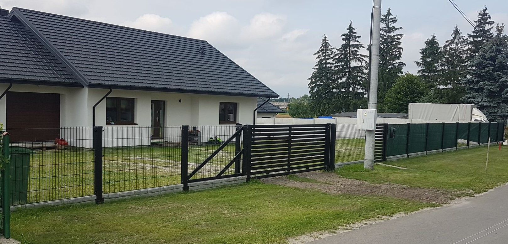 Nowoczesne ogrodzenie panelowe z bramą przesuwną w kolorze czarnym, na tle domu z ciemną dachówką i zieleni drzew.