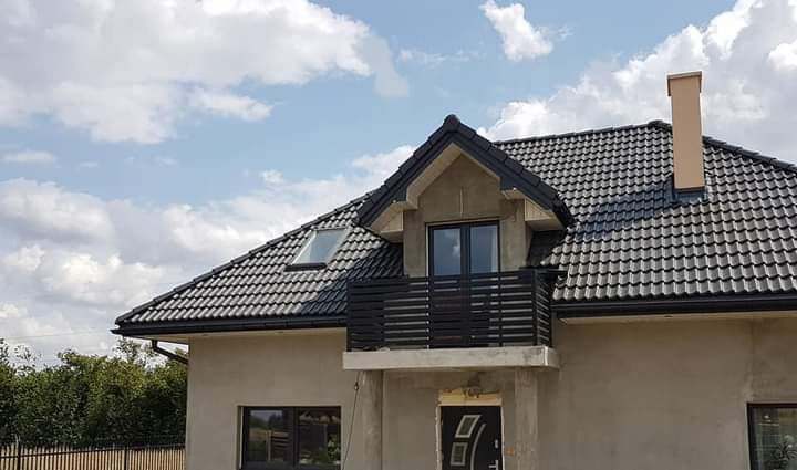 Surowy stan elewacji domu jednorodzinnego z ciemną dachówką i czarną, minimalistyczną balustradą balkonową, widok z zewnątrz w słoneczny dzień.