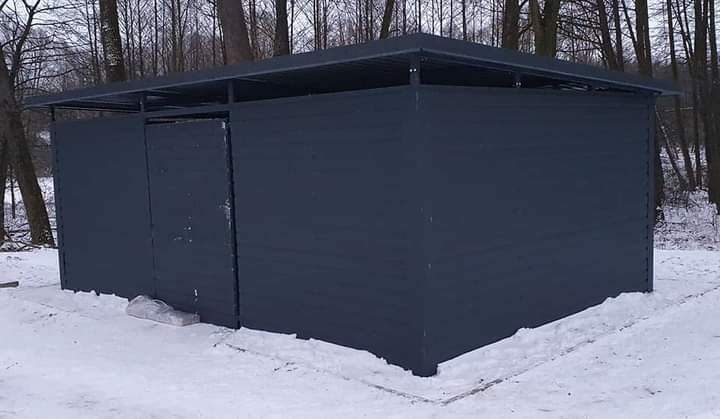 Zimowy widok na nowo wybudowaną, ciemnoszarą, metalową konstrukcję z dachem, prawdopodobnie szopę lub mały magazyn, stojącą na śniegu w lesie.