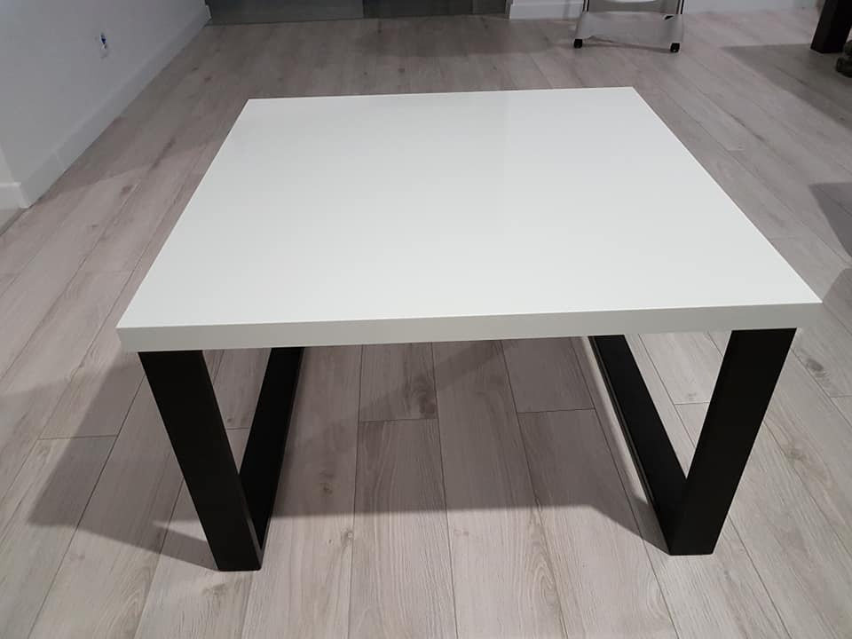 Nowoczesny stolik kawowy z białym blatem i czarnymi nogami w kształcie litery V, ustawiony na podłodze z paneli w odcieniach szarości.