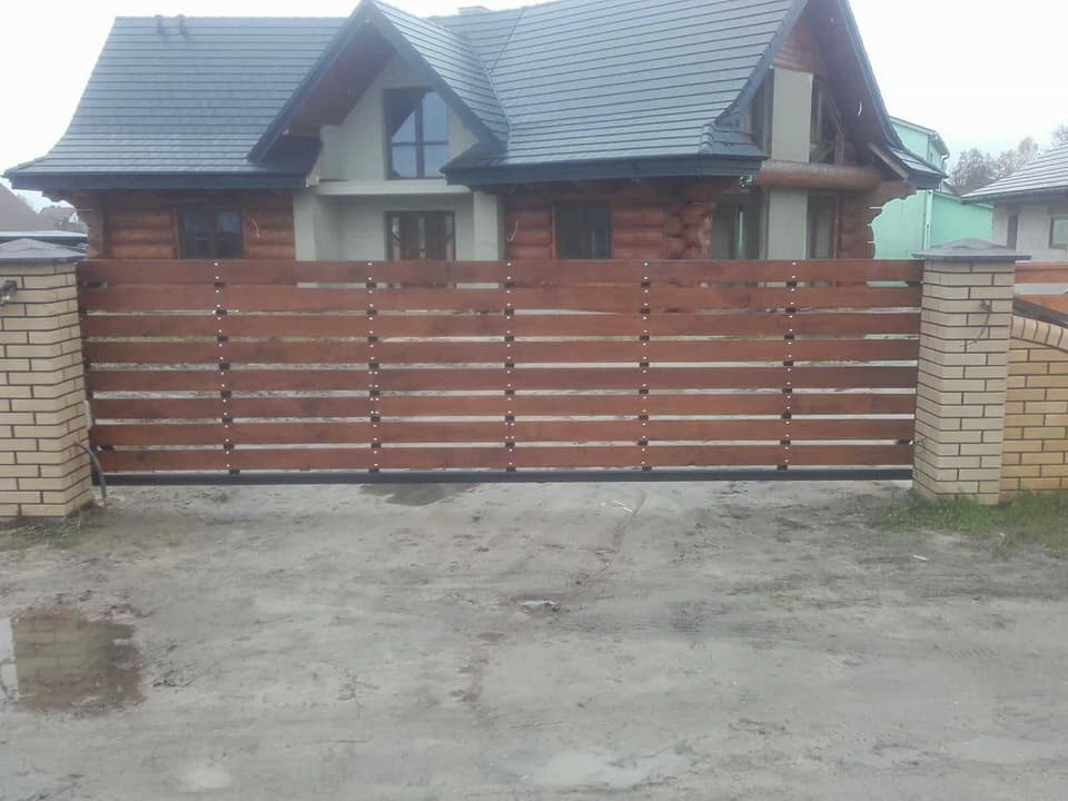 Przesuwna brama z poziomych desek w kolorze brązowym, zamontowana między murowanymi słupkami z cegły klinkierowej, na tle drewnianego domu z ciemnym dachem.