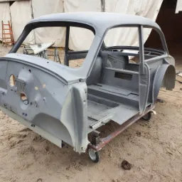 Surowa karoseria samochodu, szara, bez szyb i drzwi, osadzona na metalowym wózku na kółkach, na piaszczystym podłożu, pod namiotem.