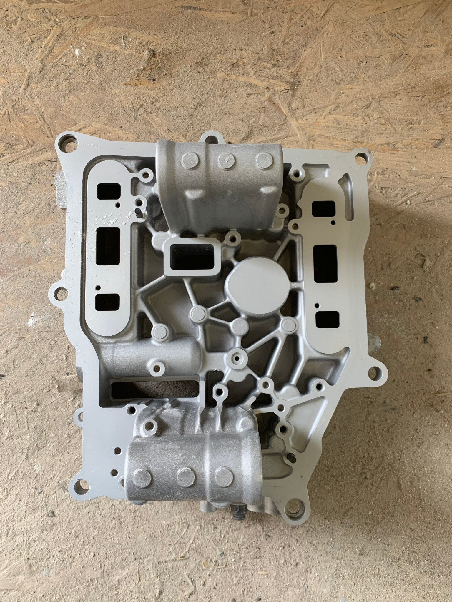 Szary, odnowiony element metalowy skrzyni biegów, leżący na drewnianej powierzchni, widoczne liczne otwory i żłobienia.