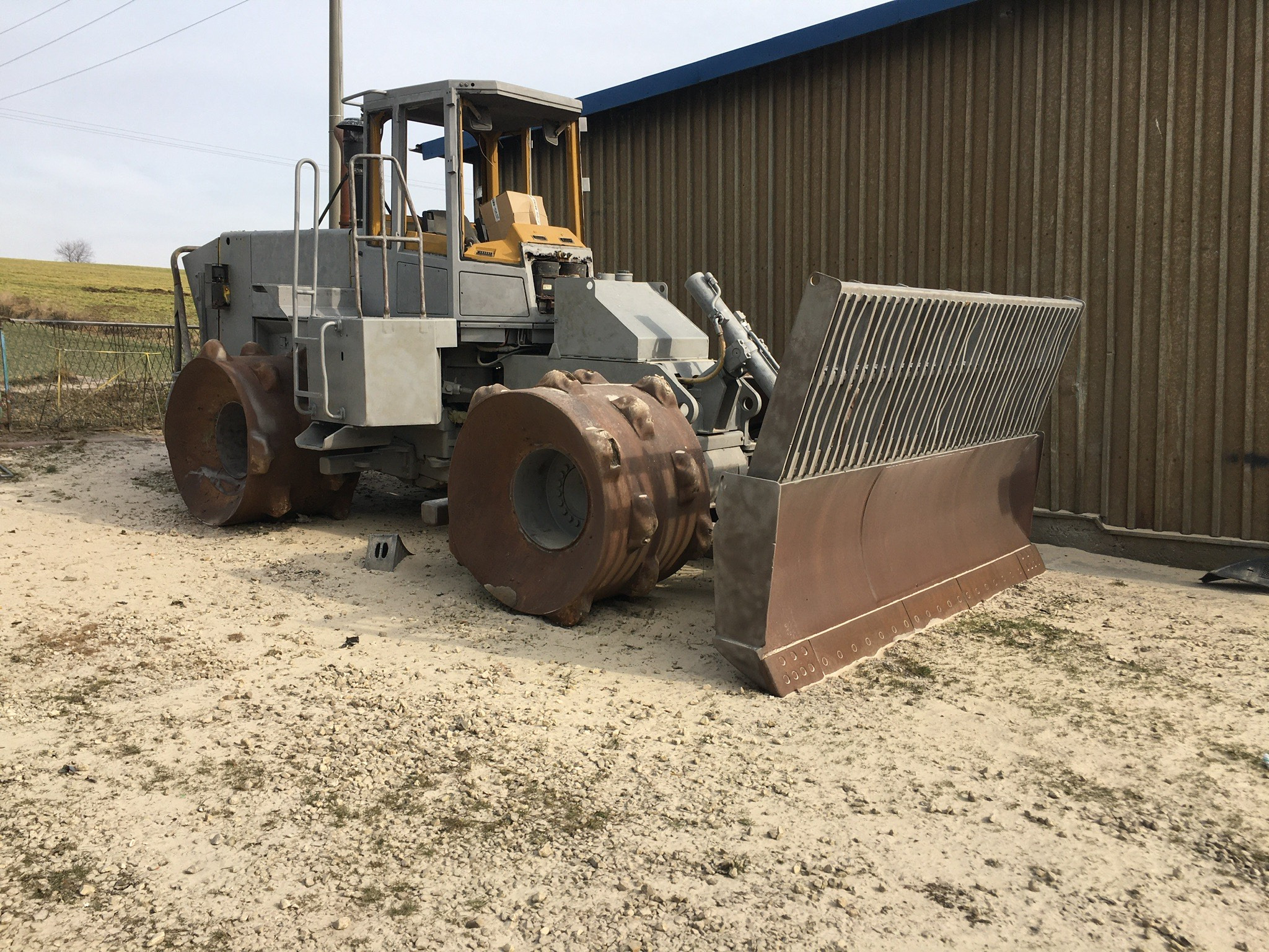 Ciężki, szary walec do ubijania gruntu z lemieszem, stojący na żwirowym podłożu przed blaszakiem.