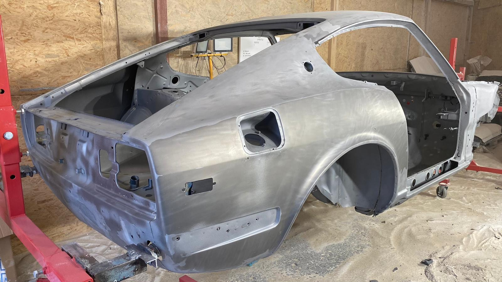 Karoseria klasycznego samochodu sportowego w trakcie renowacji, z widocznymi śladami szlifowania i przygotowania do malowania, zamocowana na czerwonym stojaku warsztatowym.