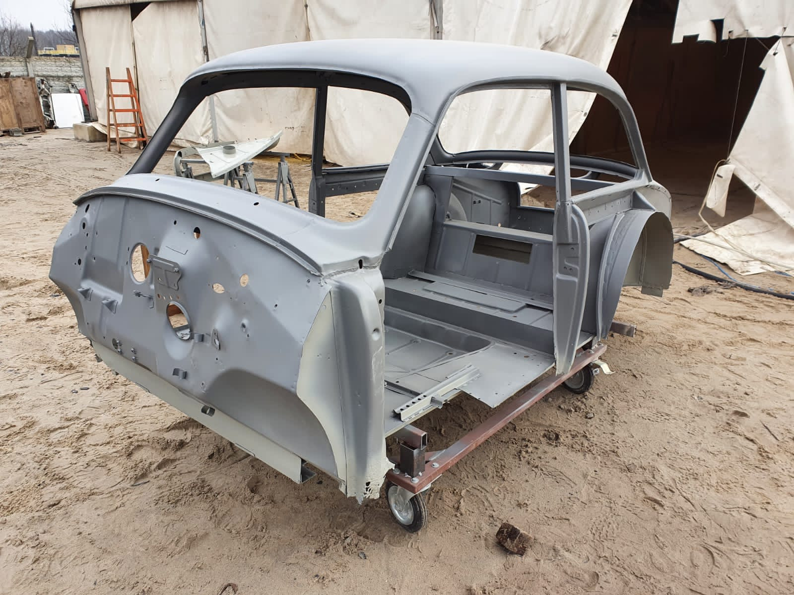Surowa karoseria samochodu, szara, bez szyb i drzwi, osadzona na metalowym wózku na kółkach, na piaszczystym podłożu, pod namiotem.
