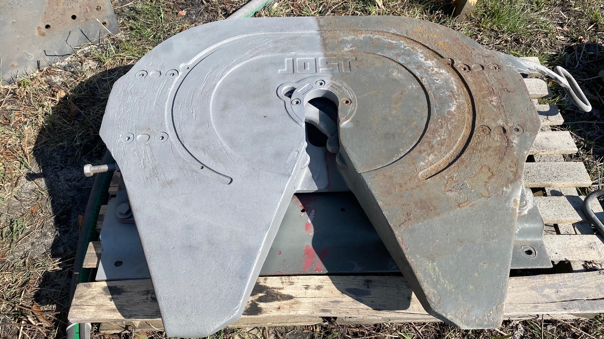 Sprzęg siodłowy Jost częściowo odnowiony, z widoczną różnicą między starą, zardzewiałą powierzchnią a nową, szarą powłoką na drewnianej palecie.