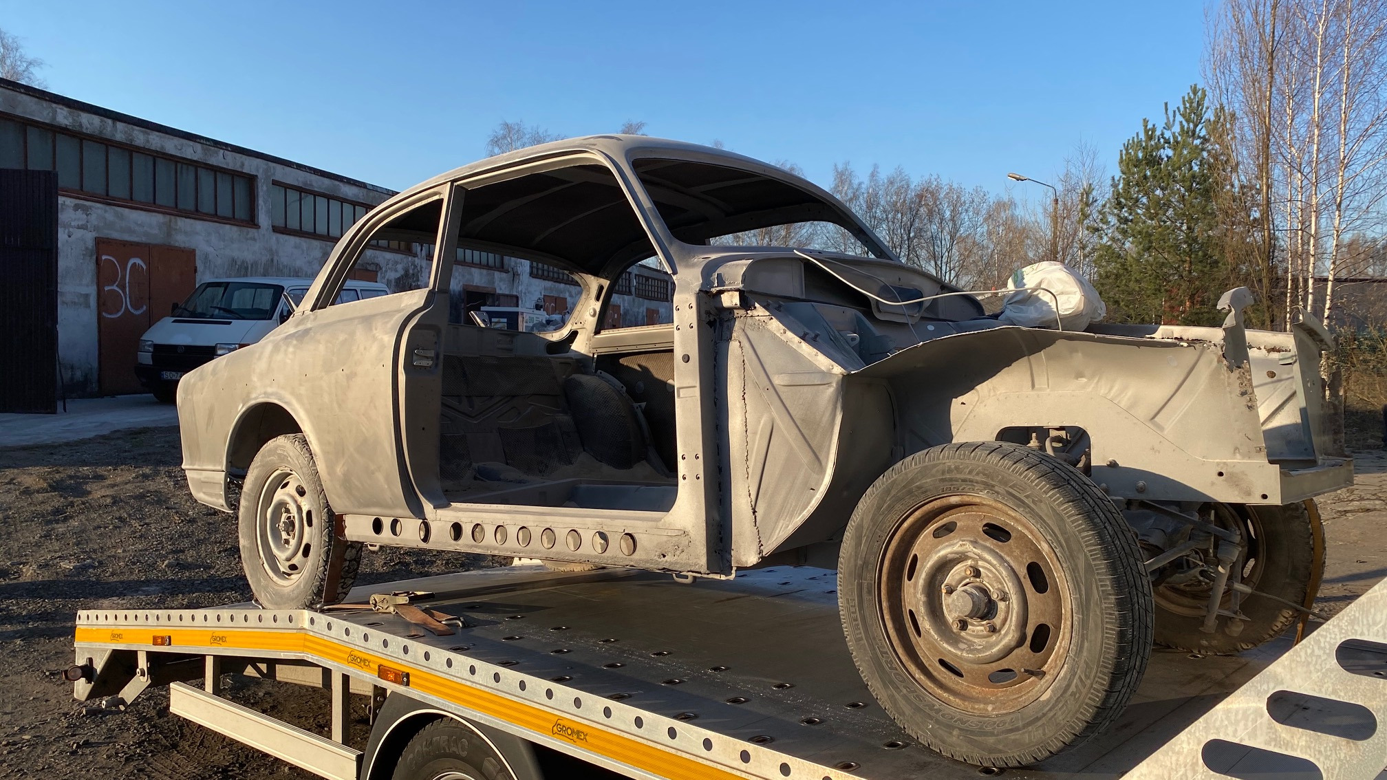 Karoseria klasycznego samochodu, pozbawiona elementów zewnętrznych, transportowana na lawecie. Widoczne ślady korozji i przygotowania do renowacji na tle budynku i drzew.