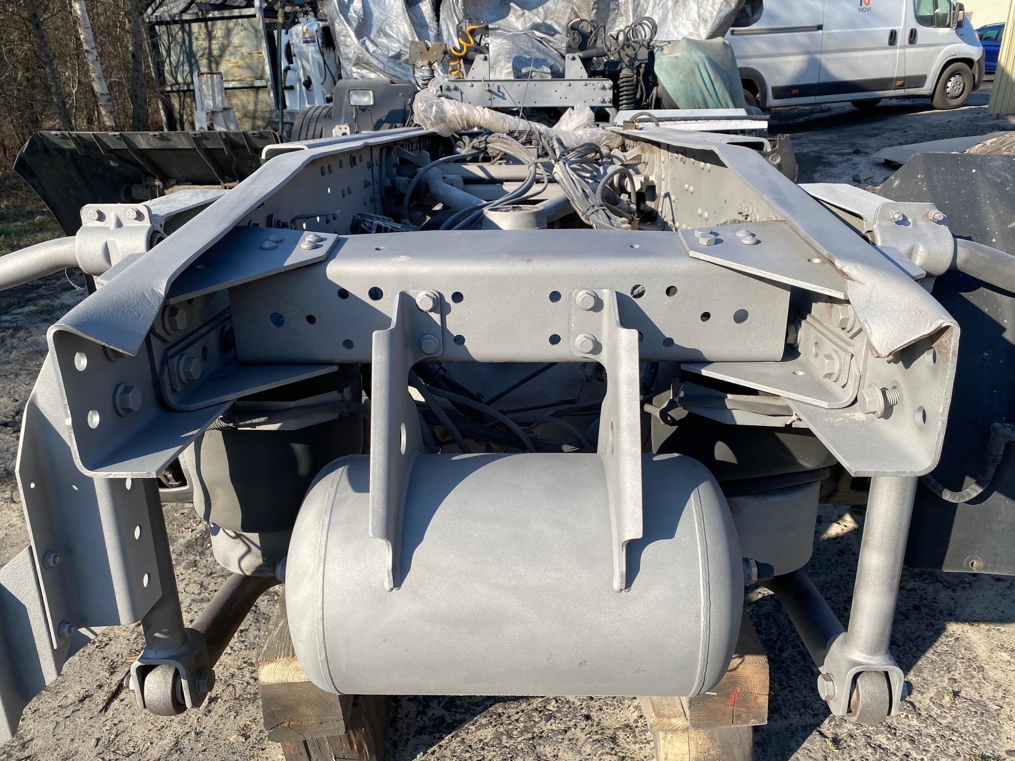 Szare, odnowione podwozie ciężarówki z widocznymi elementami zawieszenia i przewodami, stojące na drewnianych klockach, z rozproszonym światłem słonecznym.