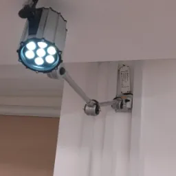 Szara lampa LED z regulowanym ramieniem zamontowana na białej ścianie, widoczne okablowanie i elementy montażowe.