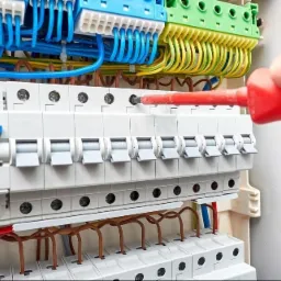 Otwarta skrzynka bezpiecznikowa z bezpiecznikami, do której dokręcana jest śrubka za pomocą czerwono-czarnego śrubokręta.