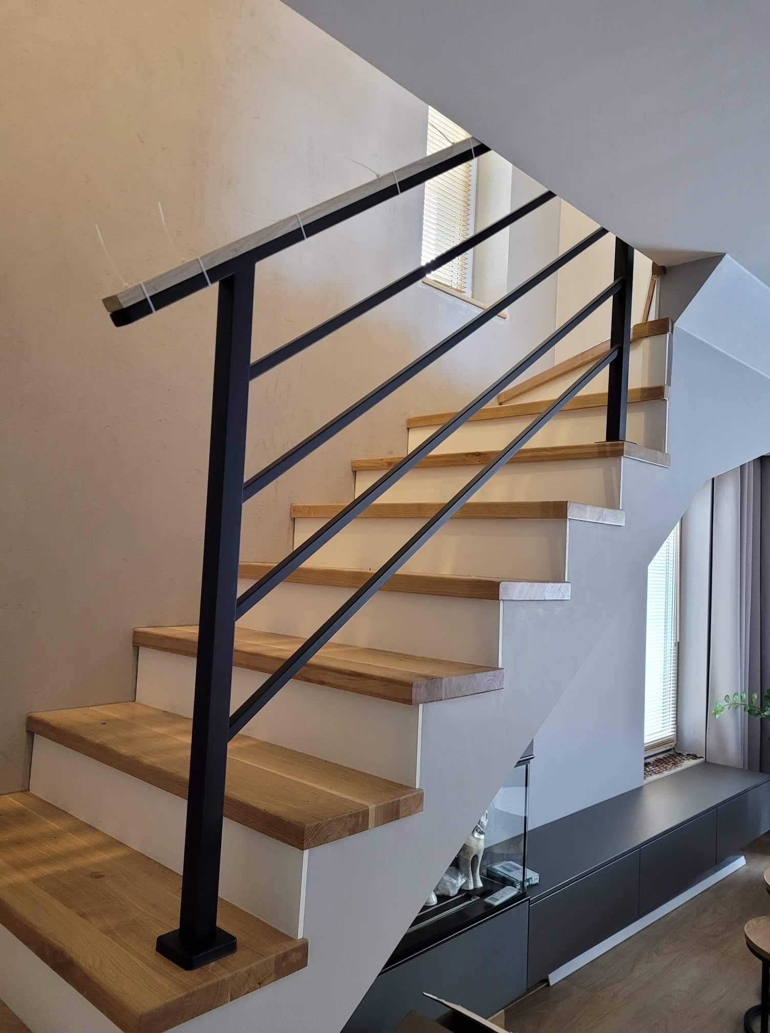 Schody z drewnianymi stopniami i czarną, minimalistyczną poręczą z rur stalowych, widoczne elementy mocujące na górnej krawędzi poręczy, jasne ściany w tle.