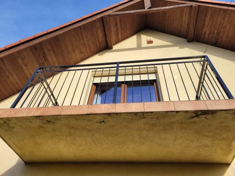 Balkon z czarną, metalową balustradą na tle żółtej elewacji domu z drewnianym podbitką dachową i brązowym oknem.