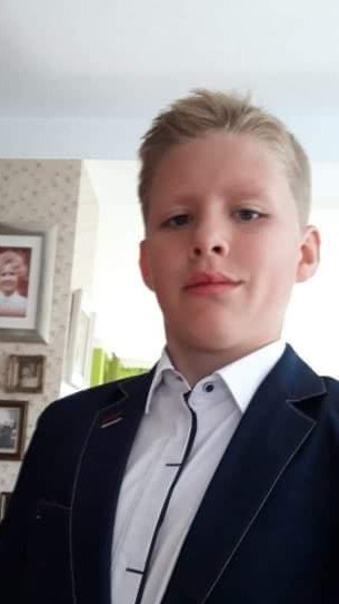 Portret młodego mężczyzny w ciemnym garniturze i białej koszuli, z jasnymi włosami, pozującego na tle ściany z tapetą i ramkami ze zdjęciami.