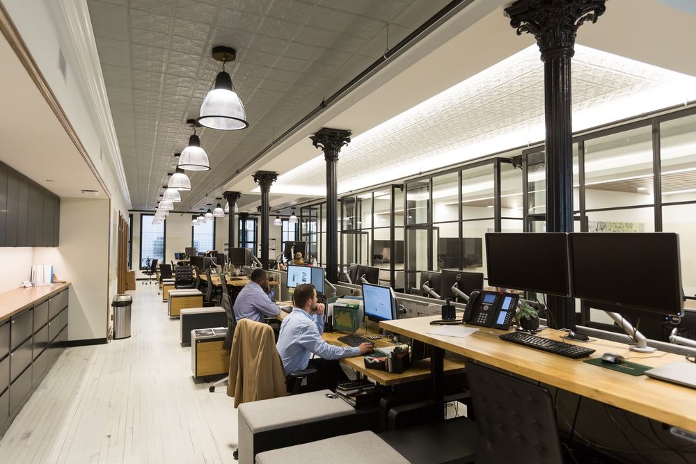 Wnętrze biura typu open space z rzędami biurek, pracownikami przy komputerach, kolumnami dekoracyjnymi i lampami wiszącymi w stylu industrialnym.