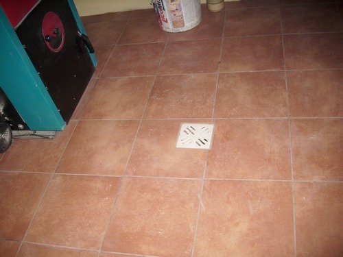 Brązowe płytki podłogowe z kratką ściekową w centralnej części, fragment pieca na paliwo stałe w lewym dolnym rogu i wiadro z zaprawą w tle.