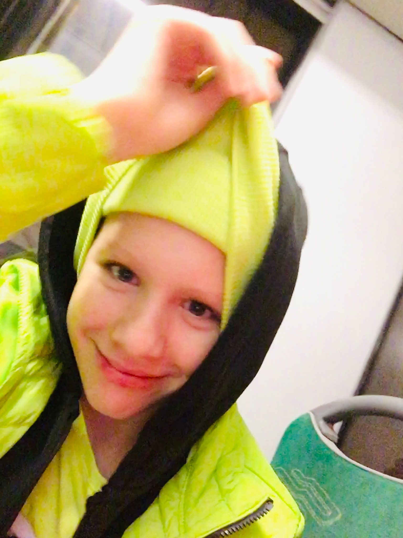 Uśmiechnięta osoba w neonowej kurtce i czarnej bluzie z kapturem, z neonową czapką na głowie, trzymająca czapkę w dłoni.