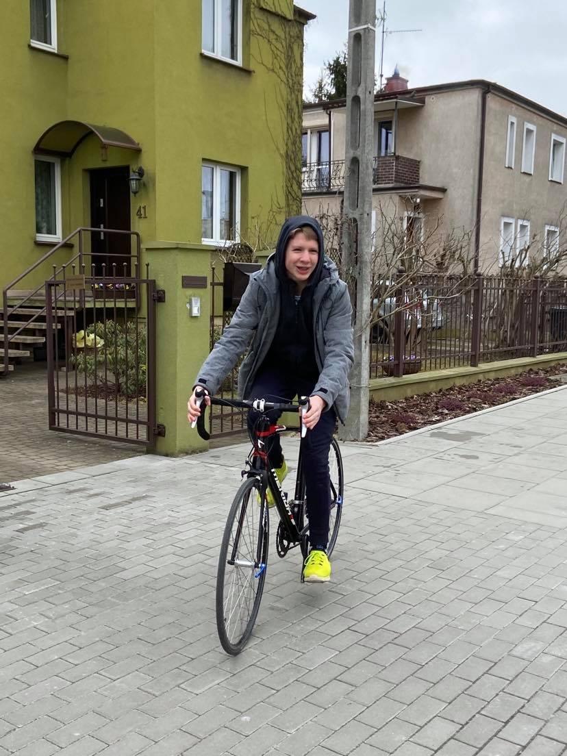 Młody chłopak w szarej kurtce z kapturem jedzie na czarnym rowerze szosowym w żółtych butach po chodniku z kostki brukowej, na tle dwóch domów i słupa energetycznego.