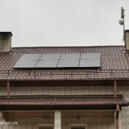 Cztery panele słoneczne zamontowane na brązowym, metalowym dachu domu jednorodzinnego z dwoma kominami i anteną satelitarną.