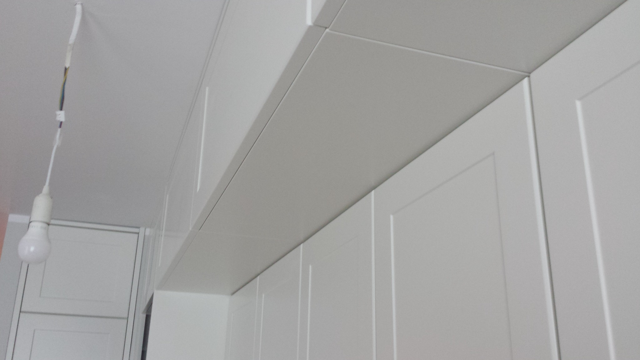 Białe, modułowe szafki kuchenne pod sufitem z widocznym kablem i żarówką w tle, ukazujące detale wykończenia i montażu.