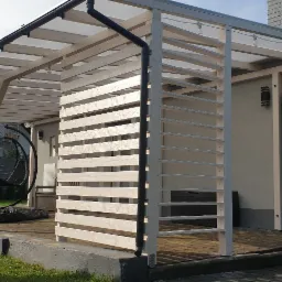 Biała pergola drewniana z przezroczystym zadaszeniem, widok z boku, z widocznym fragmentem domu i wiszącym fotelem.